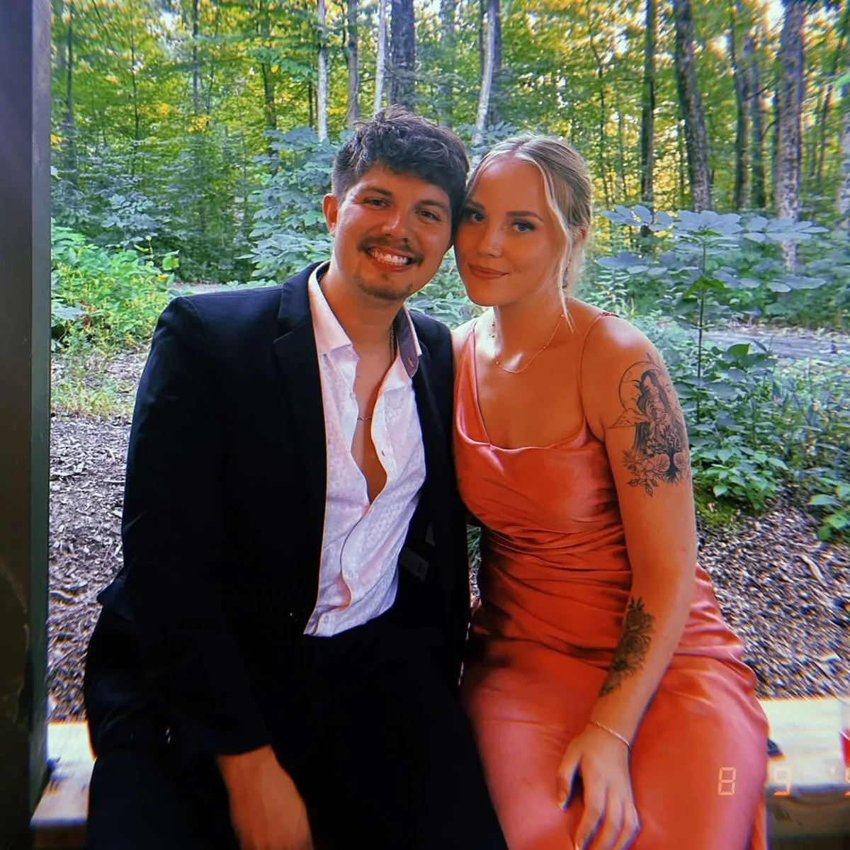 Cole Dockery in a dark jacket seated beside his partner in front of a wooded backdrop.